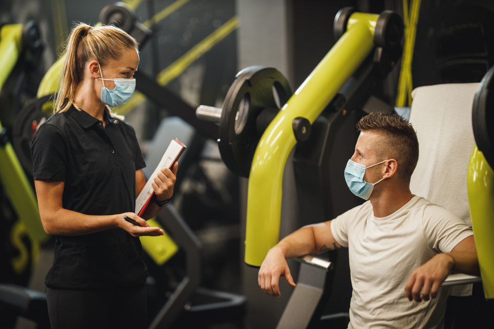 Shutterstock_1810621222 Shot of a muscular young woman with protective mask working out with personal trainer at the gym machine during Covid-19 pandemic. She is pumping up her chest muscule with heavy weight.