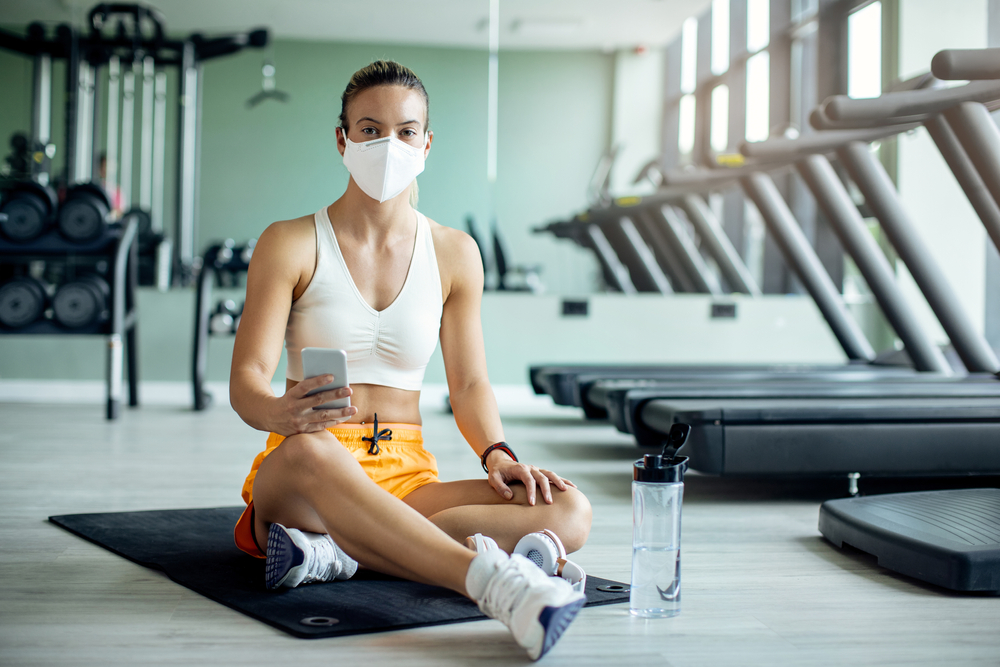 Shutterstock_1780968569 Athletic woman with face mask using smart phone while relaxing on the floor after working out in a gym during coronavirus epidemic.