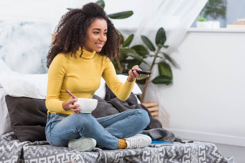 Shutterstock_ 1256409751 attractive stylish african american girl holding bowl with food and watching tv at home