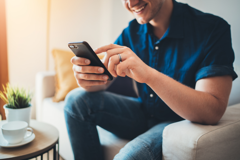 Shutterstock_749360110 Happy smiling young man using modern smartphone device while sitting on sofa at home, modern design interior, cheerful hipster guy typing an sms message at social network